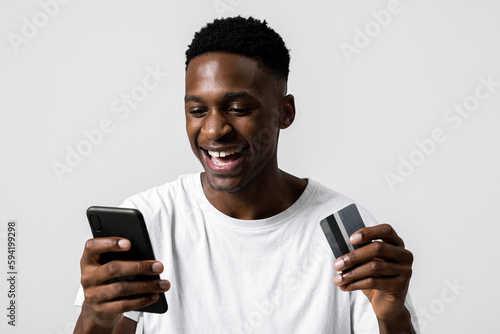 Happy delighted african american guy student received first sallary holding credit card and cellphone in hands payment process making purchase online over grey background in studio isolated. photo