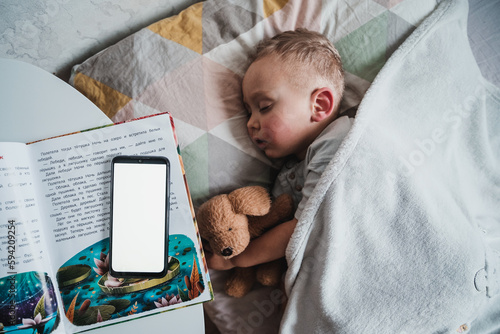 Little boy child sleeping on bed bedside table with open fairytale book and smartphone on it, kid fall asleep with bedtime phone application. Mobile app for children sleep