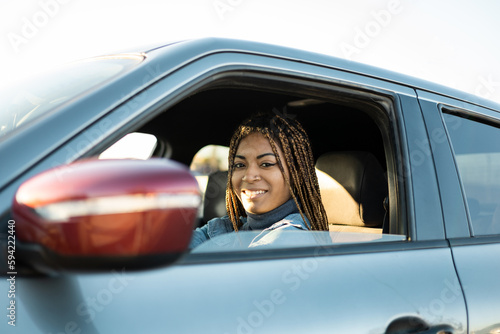 Dark-skinned young woman with braids sitting in a car looking at the camera smiling. Testing a new car, novice drivers, buying cars. Travel concept of young African women. ©  Yistocking