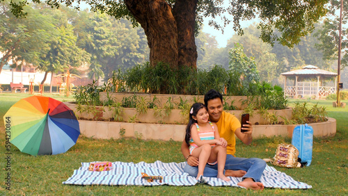 An Indian father and daughter clicking a selfie on their mobile - modern technology. Long shot of a little girl child enjoying on her dad's lap sitting under a tree in the park - family fun time to... photo