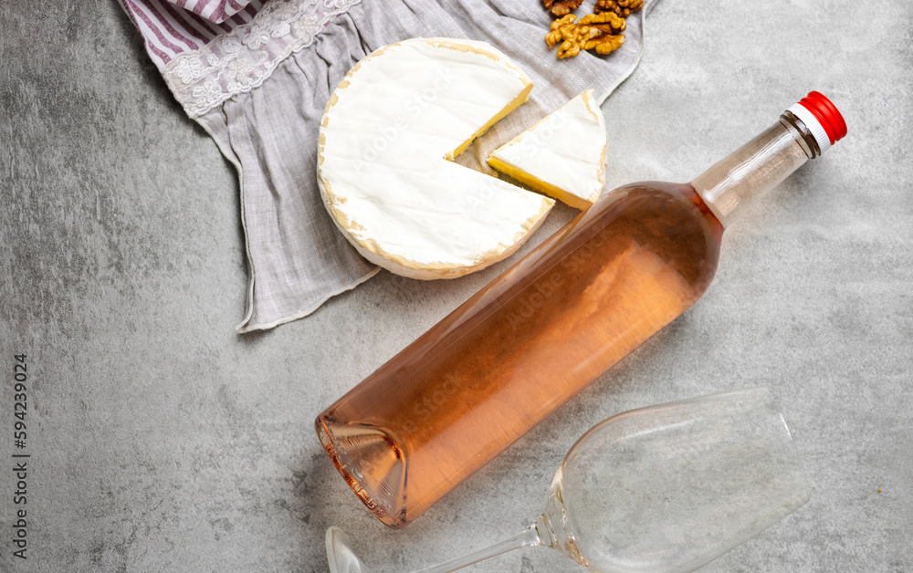 composition from rose wine bottle,glass, round cheese with one cut slice,walnuts,cork and corkscrew on kitchen striped towel or gray background tiles imitation.bottle upside down,flat lay front view