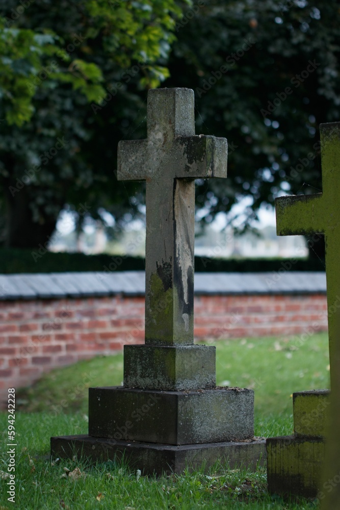 Aged stone cross in a graveyard setting, with a red brick wall in the background Aged stone cross in a graveyard setting, with a red brick wall in the background