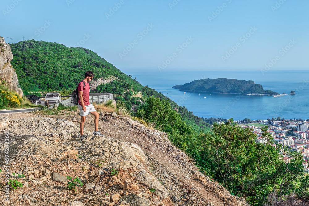 Fototapeta premium Male tourist stands on a mountain above Budva in Montenegro. Man looks down at the city from the Viewpoint on the M2.3 highway. Delightful summer landscape of mountains and sea in the Budva Riviera