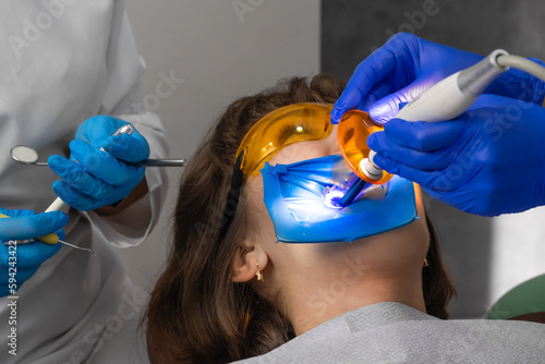 Dentist fills patient's tooth with composition of filling, and assistant shines ultraviolet lamp - to pretty woman. Tooth restoration surgery. UV for dental filling.