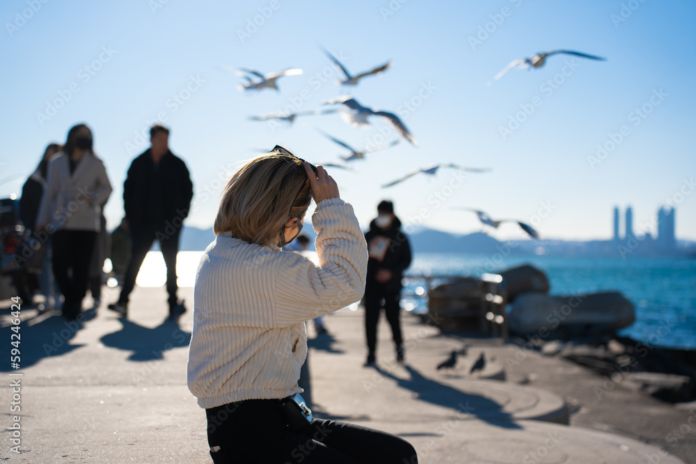 Selective focus, back side view of blond short-haired Asian woman in a corduroy jacket, sitting put sunglasses on head while admiring the sea view with many flying seagulls and tourists in background.