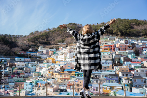 Selective focus, the back of a blond short-haired Asian woman in checks shirt standing on the hillside viewpoint with raising arms up while admiring a beautiful colorful village in Busan, South Korea. photo