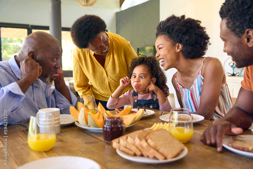 Family Shot With Grandparents Parents And Granddaughter Pulling Faces At Breakfast Around Table At Home Family Shot With Grandparents Parents And Granddaughter Pulling Faces At Breakfast Around Table At Home