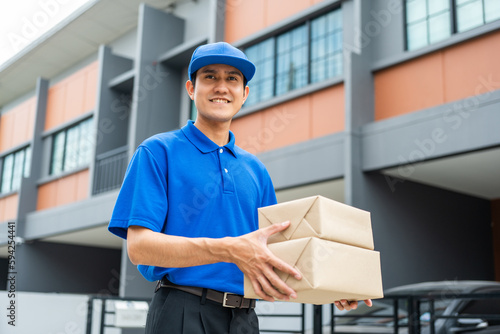 Asian delivery man with parcel in hand of blue uniform sending parcel to customer front of the house from shopping online with good service. Courier man send a package to destination. © Chanakon
