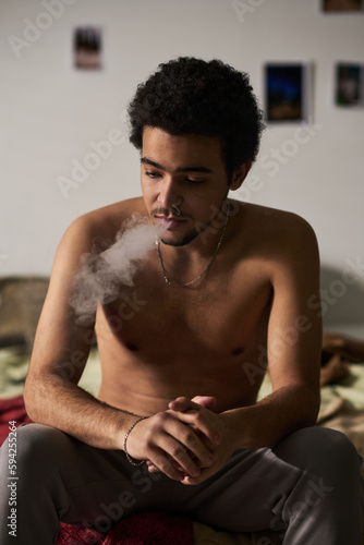 Vertical image of depressed young man smoking while sitting on bed alone in the room