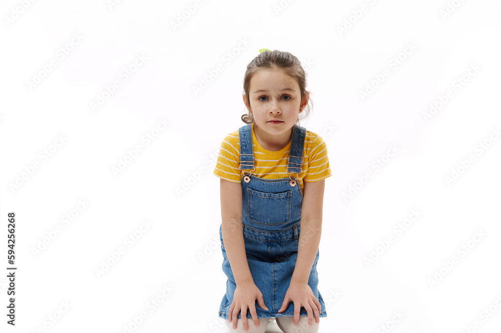 Isolated portrait on white background of adorable Caucasian preschooler child, little girl in yellow t-shirt and blue denim sundress holding hands on her knees while sitting and looking at camera Isolated portrait on white background of adorable Caucasian preschooler child, little girl in yellow t-shirt and blue denim sundress holding hands on her knees while sitting and looking at camera