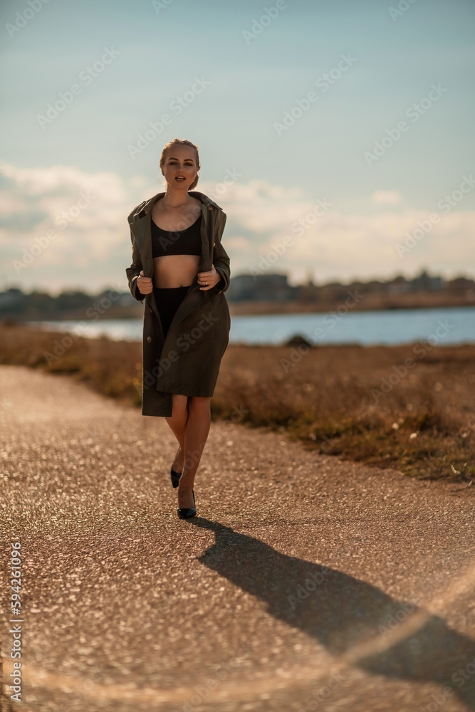 Portrait blonde sea cape. A calm young blonde in an unbuttoned khaki raincoat walks along the seashore, under a raincoat a black skirt and top