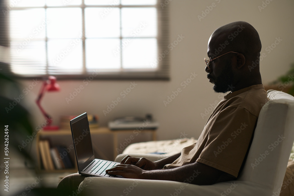 © pressmaster - African AMerican man sitting on armchair in the room and working online using his laptop