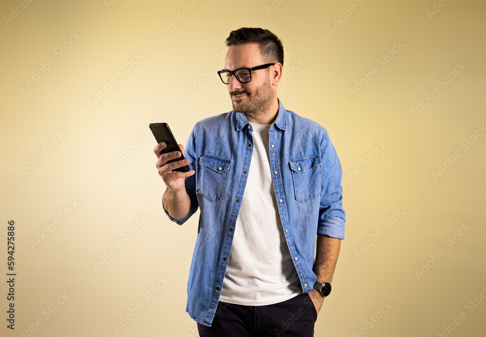 Charming male freelancer with hand in pocket checking messages on smart phone while standing isolated over beige background. He is wearing eyeglasses and blue jeans shirt Charming male freelancer with hand in pocket checking messages on smart phone while standing isolated over beige background. He is wearing eyeglasses and blue jeans shirt