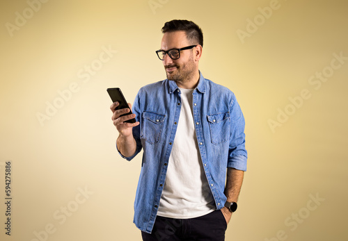 Charming male freelancer with hand in pocket checking messages on smart phone while standing isolated over beige background. He is wearing eyeglasses and blue jeans shirt © Moon Safari