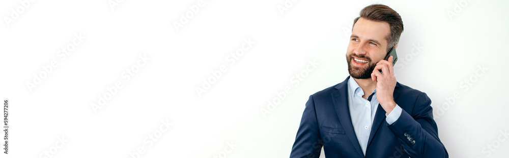 Panoramic photo of successful caucasian business man in elegant suit, stands on a white isolated background, uses smartphone, talking on the phone with a colleague, looking away, smiles. Copy-space Panoramic photo of successful caucasian business man in elegant suit, stands on a white isolated background, uses smartphone, talking on the phone with a colleague, looking away, smiles. Copy-space