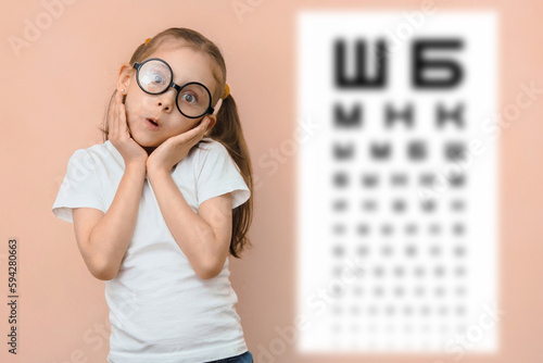 Surprised girl 5 - 7 years old in round glasses against the background of a blurred sivtsev table for checking visual acuity. photo