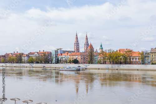 Fotografie Szeged, Hungary colorful cityscape is reflected in the river Tisza below, framed by clouds and majestic architecture on a bright day