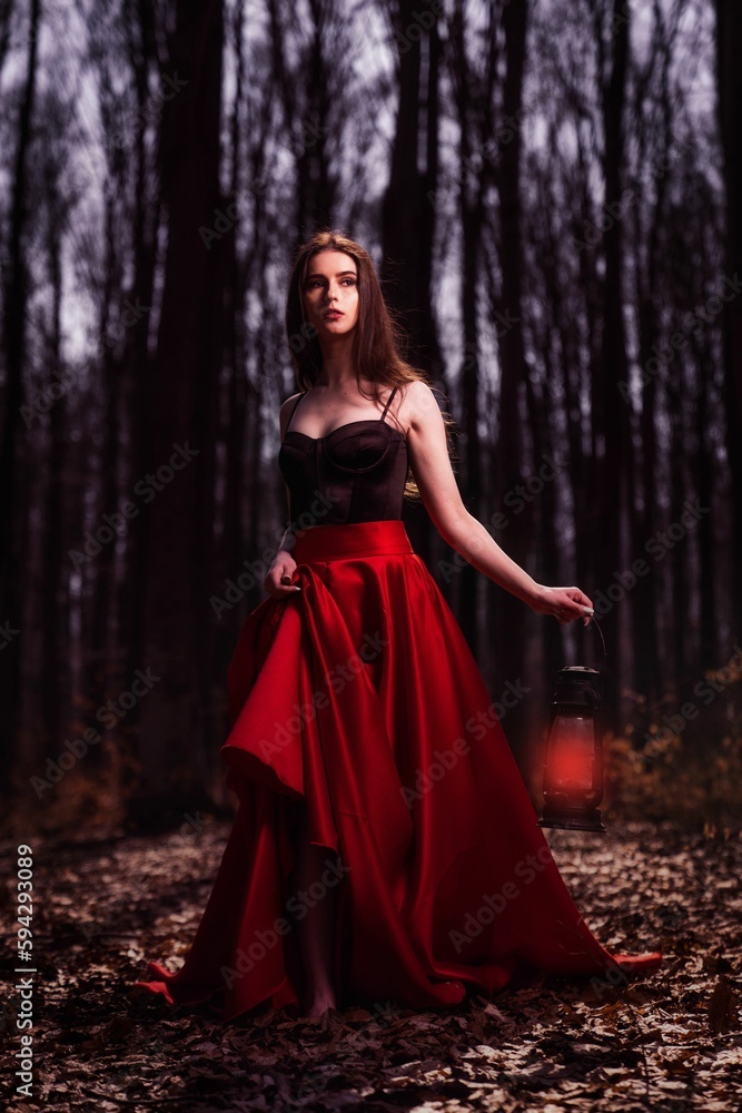Beautiful Caucasian woman in black and red dress in mysterious autumn forest with fallen dry leaves