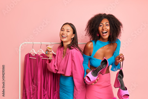 Two laughing girls in the studio, both are dressed in pink and blue clothes, black woman holds toeless shoes with heels, Asian girl holds a glass of cocktail, good shopping concept, copy space, high photo