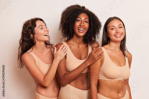 Very happy three girls wearing just beige top and underwear panties pose against white background, put their hands on shoulder of friend next to, wearing comfort concept, copy space, high quality © South House Studio