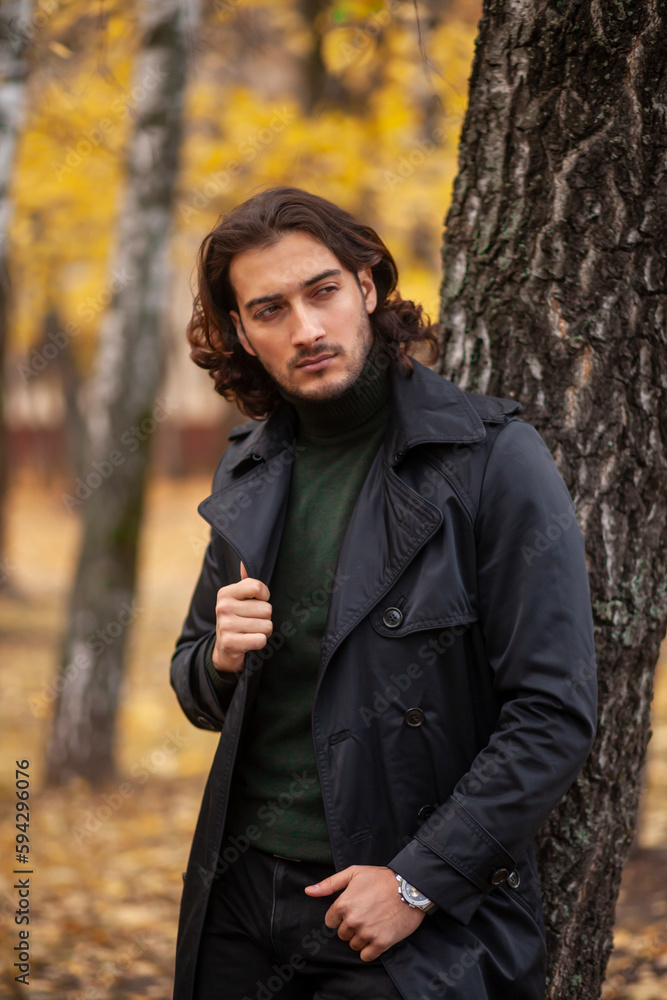 Handsome long haired man in black trench coat walking in the park in wonderful autumn day. Fashion and style concept Handsome long haired man in black trench coat walking in the park in wonderful autumn day. Fashion and style concept