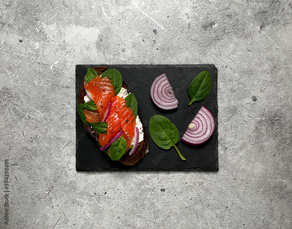 Sandwich with trout, onion rings, herbs, curd cheese on a black plate on a light background. Sandwich with trout, onion rings, herbs, curd cheese on a black plate on a light background.