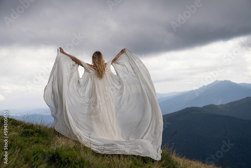 Woman in a white bridal cape standing atop a hill with a scenic landscape.