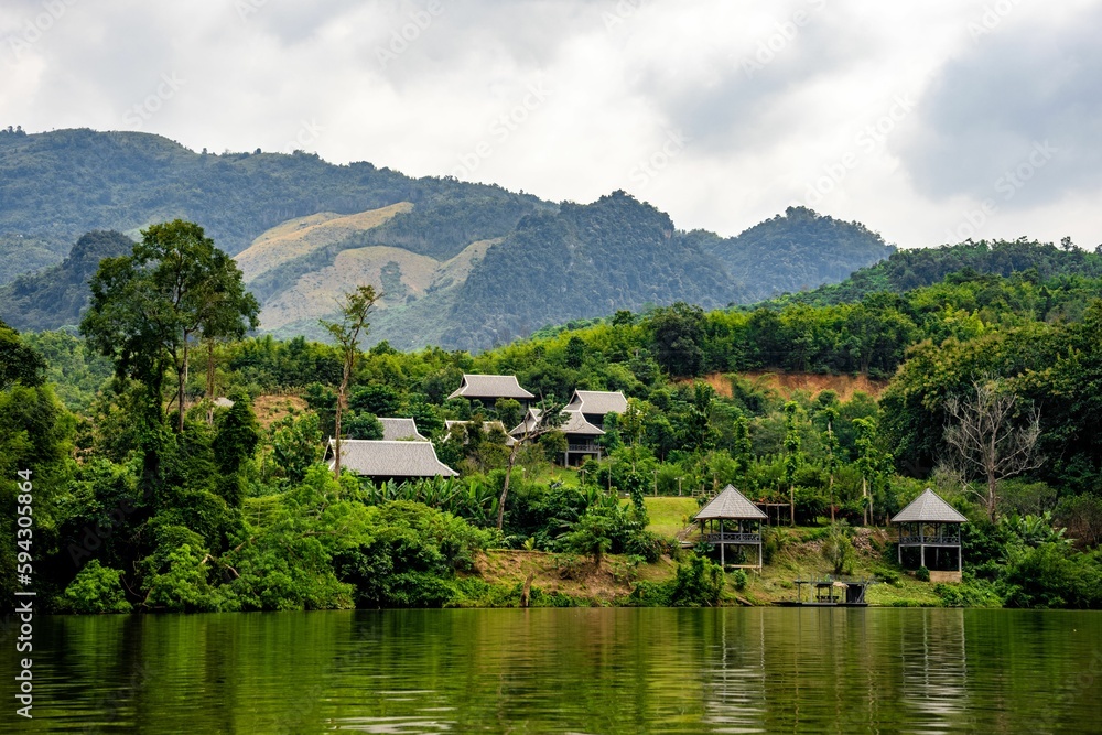 a river near some small buildings surrounded by green trees and mountain in the distance
