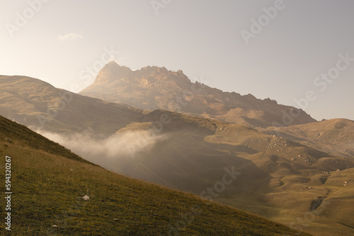 Majestic mountain landscape - high rock yellow bizarre ridge in golden sunbeams of early morning sun with white soft cloud in clear sky, mist, green hilly slopes, panorama view on valley in autumn.