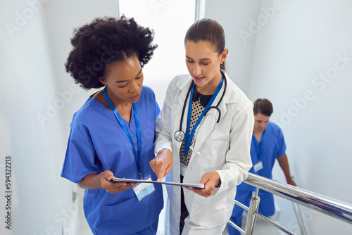 Female Doctor And Nurse Discussing Patient Notes On Stairs In Hospital Building