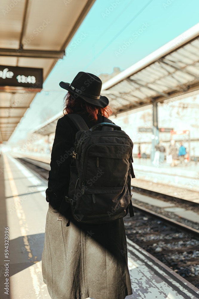 Fototapeta premium Vertical back view of a young female wearing black coat and backpack at the station