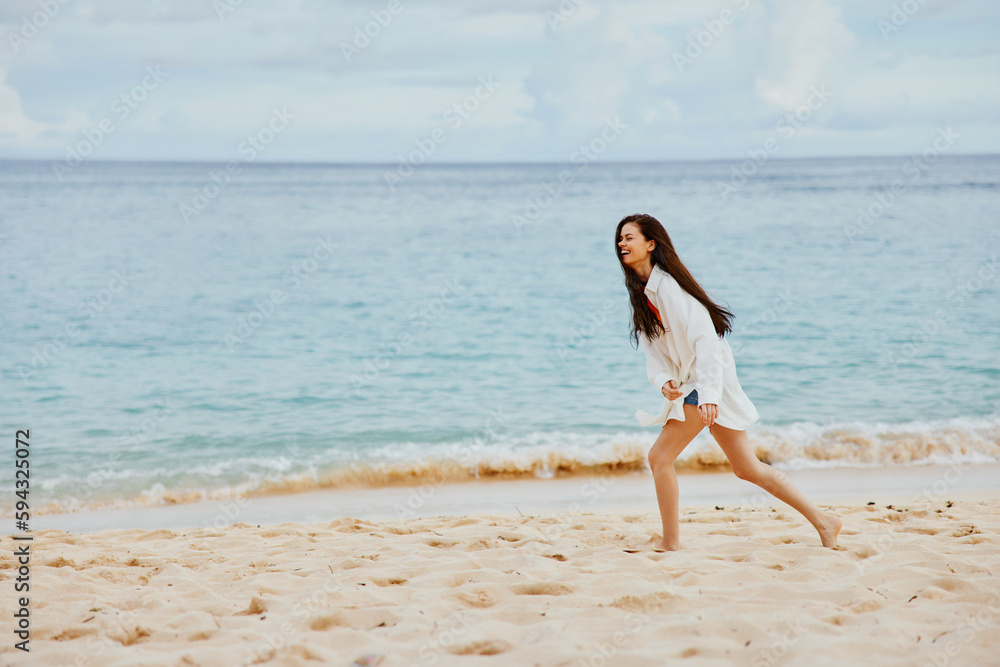 Sports woman runs along the beach in summer clothes on the sand in a yellow T-shirt and denim shorts white shirt flying hair ocean view, beach vacation
