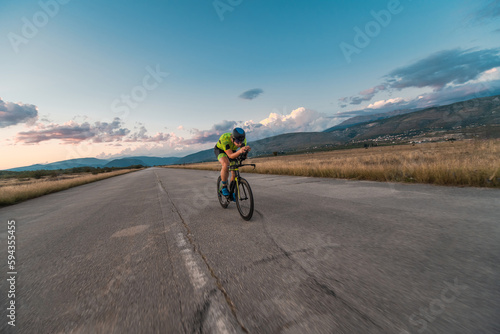  Triathlete riding his bicycle during sunset, preparing for a marathon. The warm colors of the sky provide a beautiful backdrop for his determined and focused effort. © .shock