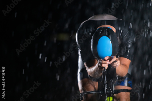 Fototapeta Naklejka Na Ścianę i Meble -  A triathlete braving the rain as he cycles through the night, preparing himself for the upcoming marathon. The blurred raindrops in the foreground and the dark, moody atmosphere in the background add