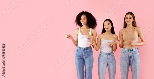 Look there. Three happy diverse women pointing fingers aside at free space, advertising something on pink background