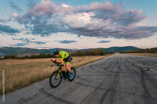  Triathlete riding his bicycle during sunset  preparing for a marathon. The warm colors of the sky provide a beautiful backdrop for his determined and focused effort.