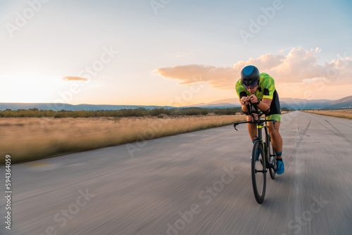  Triathlete riding his bicycle during sunset, preparing for a marathon. The warm colors of the sky provide a beautiful backdrop for his determined and focused effort.