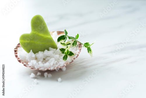 Jade green Gua sha and two small green branches on the sea shell full of sea salt on the light marble background. Copy space. Spa composition