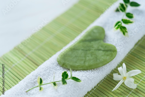 Green jade Gua sha and two small branches of young leaves on the white towel on the green bamboo mat with white small flower. Flat lay. Spa composition. Freshness 