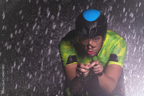 Fototapeta Naklejka Na Ścianę i Meble -  A triathlete braving the rain as he cycles through the night, preparing himself for the upcoming marathon. The blurred raindrops in the foreground and the dark, moody atmosphere in the background add