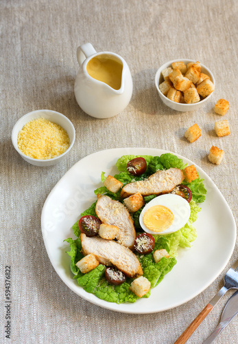 Caesar salad with croutons, fried chicken, cherry tomatoes, egg and parmesan cheese on a light plate with a fork, on a linen tablecloth in a plate of grated parmesan cheese and cherry tomatoes