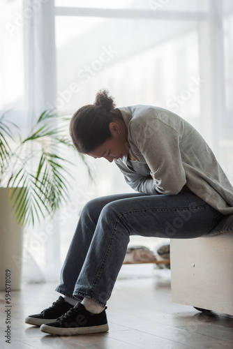 Sad multiracial woman feeling unwell while sitting on bed at home. © LIGHTFIELD STUDIOS