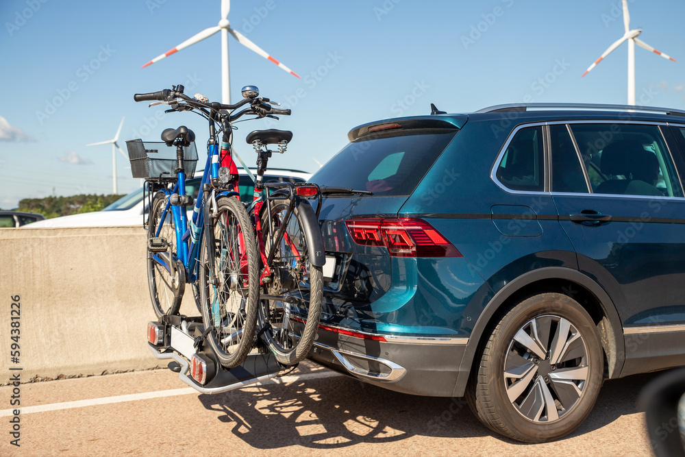 Fototapeta premium Back view modern white family wagon car with mounted roof box trunk and bike tail carrier driving european highway road against blue sky summer day. Lifestyle travel adventure trip journey concept