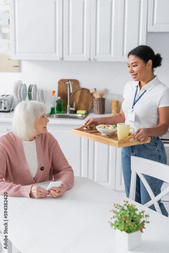 cheerful multiracial social worker holding tray with lunch for senior woman with grey hair. cheerful multiracial social worker holding tray with lunch for senior woman with grey hair.