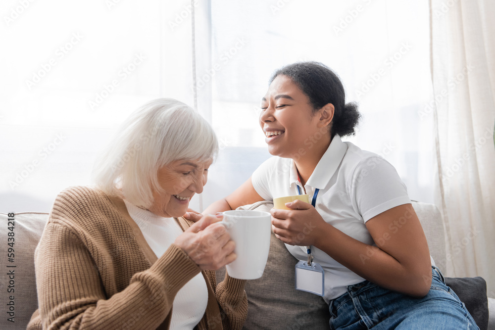 cheerful multiracial social worker laughing with senior woman while having tea in living room. cheerful multiracial social worker laughing with senior woman while having tea in living room.