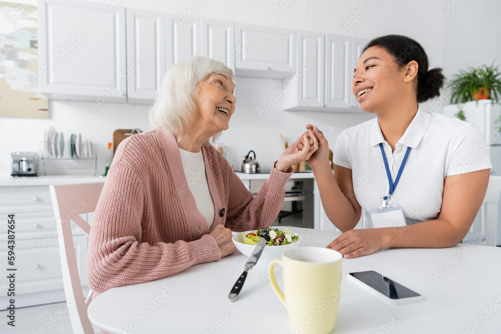 happy retired woman with grey hair holding hands with cheerful multiracial social worker next to lunch on table. happy retired woman with grey hair holding hands with cheerful multiracial social worker next to lunch on table.