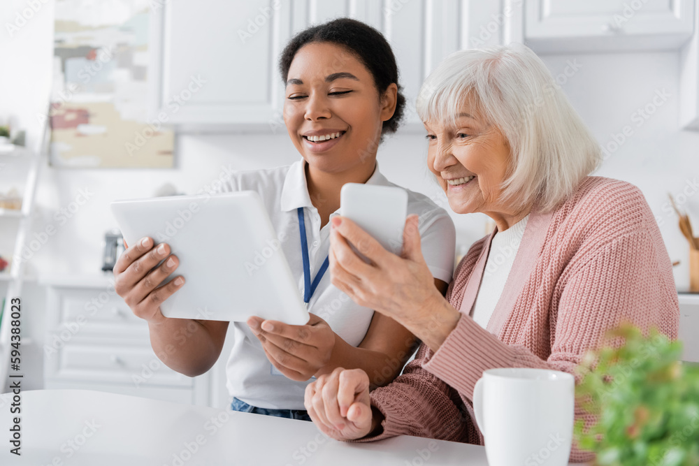happy multiracial social worker holding digital tablet near senior woman with smartphone in kitchen. happy multiracial social worker holding digital tablet near senior woman with smartphone in kitchen.