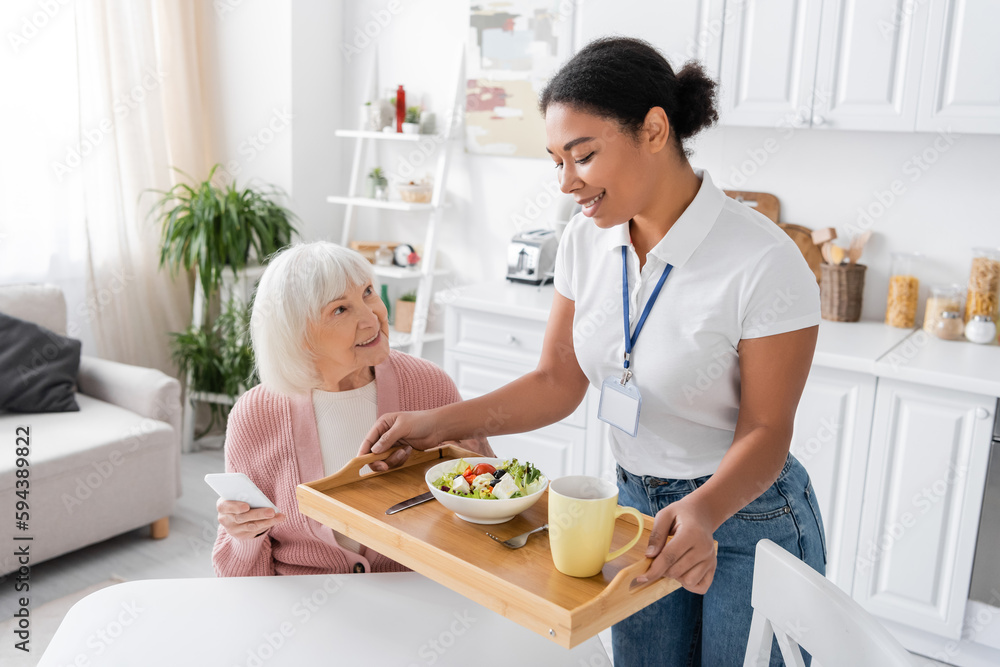 happy multiracial social worker holding tray with lunch for senior woman with grey hair.