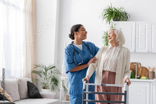 Fotografía happy multiracial nurse in uniform laughing with retired woman while standing near walking frame at home