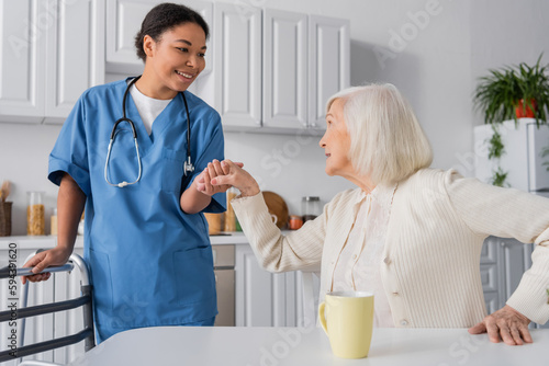 happy multiracial nurse in blue uniform holding hand of senior woman with grey hair while standing near walker.